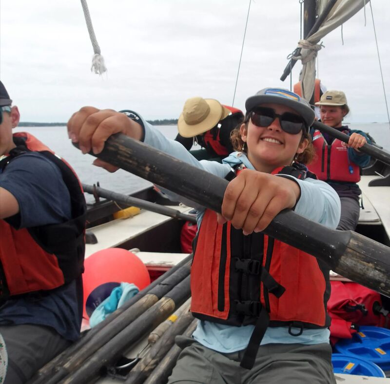 A group of people are on a boat, possibly a sailboat, wearing life jackets. The main focus is on a woman in the foreground who is smiling and holding a large wooden pole, likely part of the boat's rigging or steering mechanism. Other people are visible in the background, also wearing life jackets, and the boat appears to be on the water with land visible in the distance.